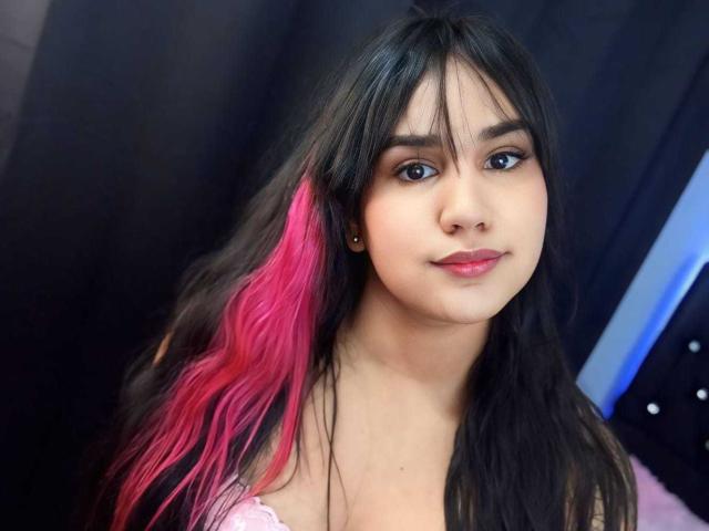 KimZenith with long black hair and pink highlights, blue eyes, and pink lipstick, smiling at the camera, wearing a pink lace top, showing her tits subtly.