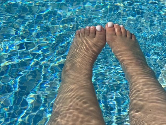 Barbarica's feet in a clear blue pool, legs submerged, toes with white nail polish, sunlight creating wavy water patterns on her skin.