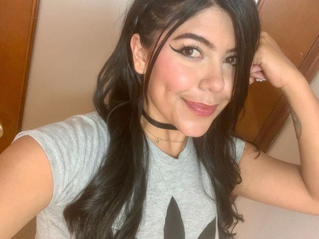 CatalinaReiyna with long black hair, wearing a gray tee, black choker, and winged eyeliner, smiling at the camera, hand in her hair, showing her cute, pink-cheeked face.