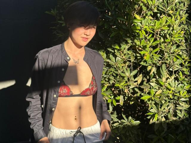 LissyLuxe met kort zwart haar, die een rode bh met bloemenmotief draagt en een open zwart shirt, grijze broek, kleine borsten, een plat buikje en een piercing in haar navel, staand in het zonlicht..