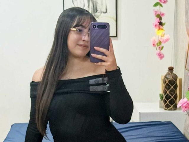 Ann Fisher, avec de longs cheveux bruns, des lunettes et un haut noir à une épaule, tenant un téléphone violet, prenant un selfie devant un miroir, montrant ses seins, souriante, dans une chambre..