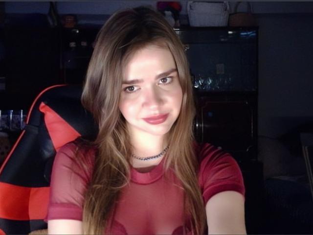 SelmaOtinger with long brown hair, red sheer top, and a black choker, smiling at the camera, showing her small tits, in a dimly lit room with a red and black chair.