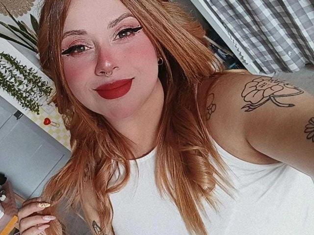 PauleetteRoux heeft rood haar, een wit tanktopje, rode lippenstift, een neusring, tatoeages en zwarte eyeliner. Ze glimlacht met een licht omhoog gekeerd neusje en houdt een potlood vast..