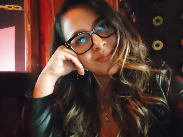 LiaDiane, com olhos azuis, óculos pretos, cabelo castanho ondulado e um leve sorriso, com a cabeça apoiada na mão, vestindo uma jaqueta de couro preta. Seus seios estão parcialmente visíveis..