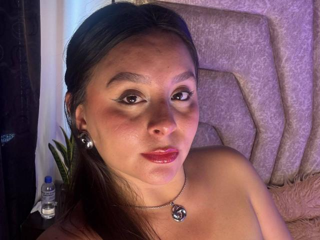 EmaJames, avec des cheveux bruns, du rouge à lèvres rose et un collier, portant des boucles d'oreilles argentées, regardant la caméra, nue sur le torse, avec une bouteille sur le côté gauche..
