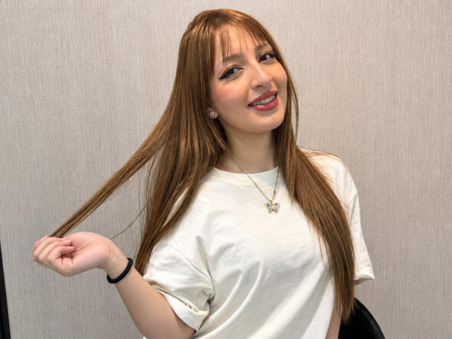 MiluSantoro mit langen, braunen Haaren, weißem T-Shirt, lächelnd, die Haare haltend, rosa Lippenstift, kleine Ohrringe, schwarzes Armband, Halskette, helle Haut, stehend, einfacher Hintergrund, süß, lässig, entspannt..