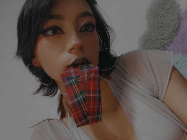 HenutmireRossi with black hair, wearing a grey shirt, holding a red plaid cloth in her mouth, looking at the camera with black eyeliner. Her tits are slightly visible.