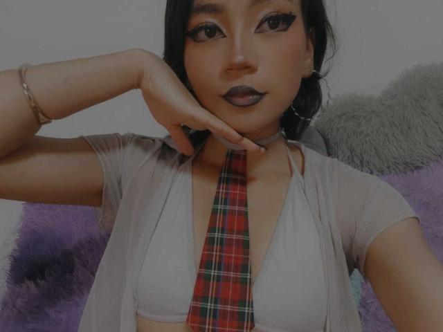 HenutmireRossi with dark lipstick, wearing a sheer top, plaid tie, and bracelet, touching her chin, showing her tits, and looking at the camera.