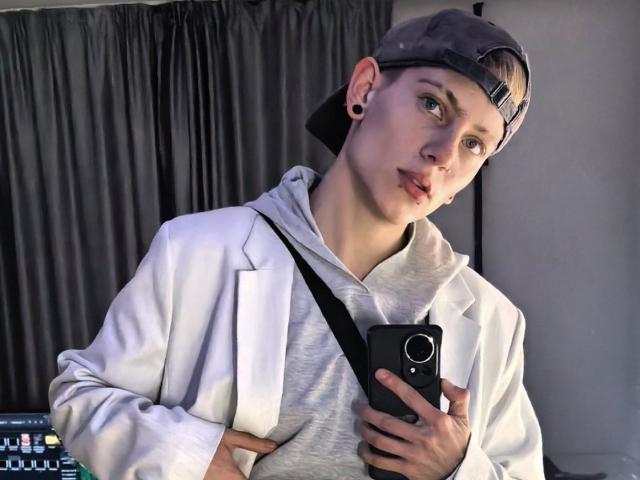 Une jeune femme au teint clair, portant un sweat à capuche gris, une veste blanche et une casquette à l'envers, tenant un téléphone, regardant de côté avec des lèvres boudeuses, avec des piercings aux oreilles noirs et un anneau au nez..