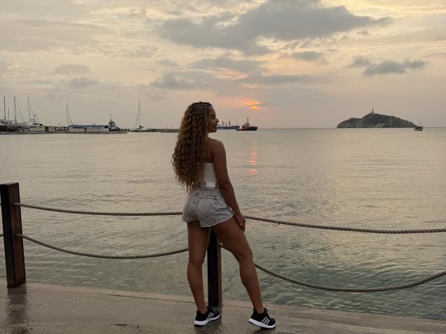 Emma, eine Lateinerin mit langen, lockigen Haaren, einem kleinen weißen Oberteil, grauen Shorts und schwarzen Turnschuhen, steht auf einem Steg und blickt auf einen ruhigen Sonnenuntergang über einem Ufer mit Booten und einer kleinen Insel..