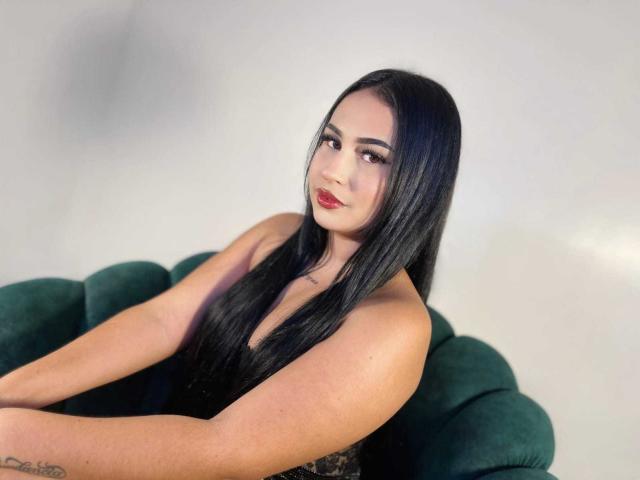 HannahBecker with long black hair, red lipstick, and a tattoo on her arm, sitting on a green velvet chair, looking at the camera with a slight smile.