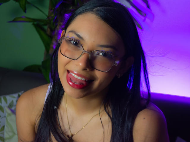 ValeryDoshel with black hair, glasses, red lipstick, and gold necklace, tongue out, smiling, showing her ass, sitting against a green and purple lit background.