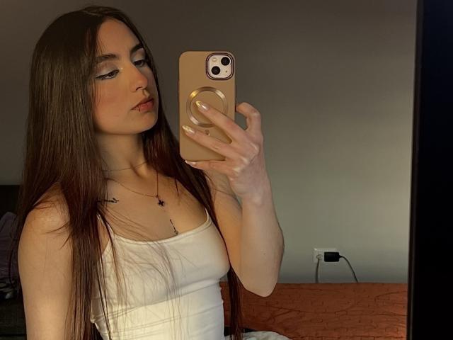 MiaHarperr, con lunghi capelli castani, canotta bianca, che tiene il telefono per un selfie allo specchio, mostrando il seno, con un leggero rossore sulle guance, una collana dorata, in una stanza poco illuminata..