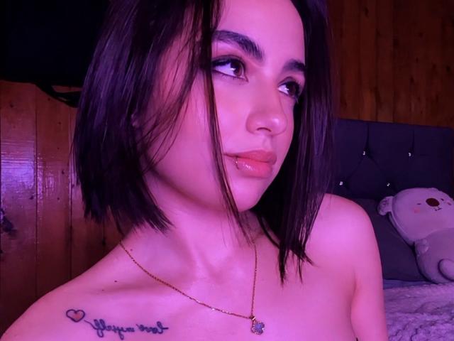 MiaHarperr, con la pelle leggermente viola, labbra lucide e un caschetto nero, indossa una collana, mostrando un tatuaggio sulla spalla, e guarda di lato. Seno scoperto, senza abiti visibili..