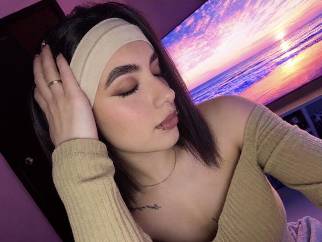 MiaHarperr, avec ses cheveux foncés, son bandeau beige et son haut côtelé à épaules dénudées, se touche la tête, montre son piercing aux lèvres et son tatouage, dans une pièce éclairée en violet avec un écran de télévision diffusant un coucher de soleil..