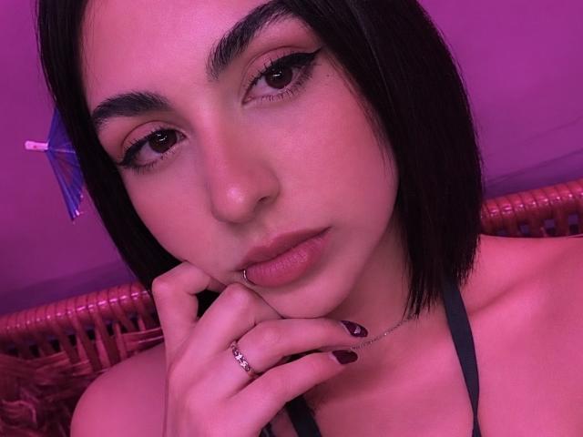MiaHarperr a une peau rosée-violette, une coupe de cheveux courte noire, du khôl, du rouge à lèvres rouge foncé, une lèvre inférieure percée, une bague au doigt, des ongles rouges foncés, un collier ras du cou, et elle fait un clin d'œil à la caméra..