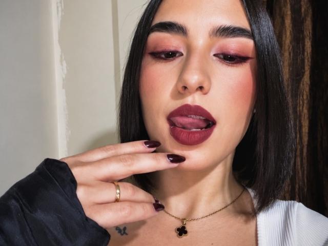 MiaHarperr heeft donkerrode lipstick, roze oogschaduw en zwarte nagels. Ze raakt haar lippen aan met haar vingers, draagt een gouden ketting en een zwarte jas. Haar tong steekt een beetje uit..