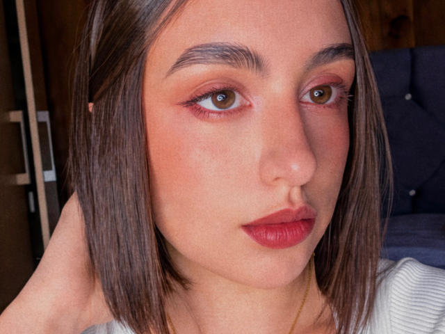 Das Gesicht von MiaHarperr, mit ihren braunen Augen, rosafarbenem Lidschatten, rotem Lippenstift und glattem, braunem Haar, mit einem leichten Lächeln, trägt ein weißes Oberteil und blickt in die Kamera..