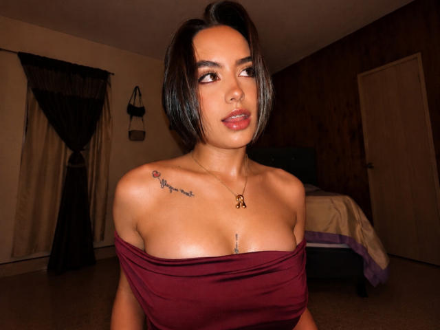 MiaHarperr, avec ses cheveux noirs courts, ses yeux marrons et son rouge à lèvres rouge, porte un haut bordeaux à une épaule, laissant entrevoir de petites seins avec des tatouages sur son épaule droite et sa poitrine..