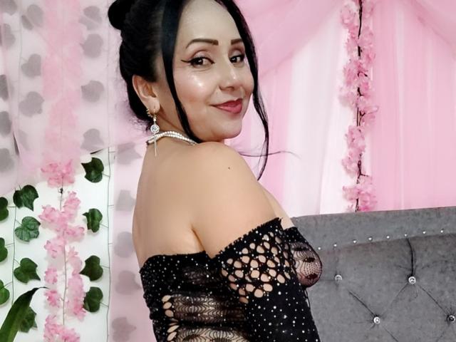 Zaria69 with black hair in a bun, wearing a sparkly black off-shoulder top, exposing her tits, smiling at the camera, pink floral background, and grey tufted couch.