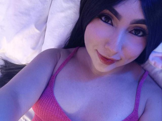 ChloeMonet with long black hair, wearing a pink ribbed bra, smiling at the camera, showing her tits, lying on white pillows, blue-purple lighting.