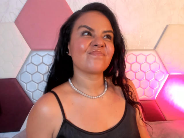 ArielleWilde with long black hair, smiling slightly, wearing a black spaghetti strap top and pearl necklace, looking to the side. She's got a cute, chubby face.