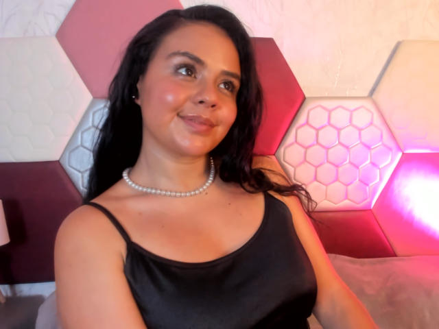 ArielleWilde with long black hair, wearing a black satin top, pearl necklace, and smiling. She's got medium tits, light brown skin, and a cute face. She's sitting on a bed.