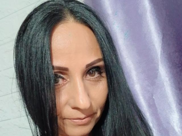 RoxiyG a de longs cheveux noirs, des yeux marrons et un léger sourire, avec un maquillage discret. Son visage est centré, révélant une peau lisse et un aspect naturel..