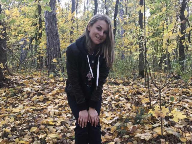 BelleLisaG, avec des cheveux blonds, souriante, portant une veste noire et un sweat à capuche, debout dans une forêt avec des feuilles jaunes, les mains sur les genoux, ambiance automnale, mignonne, lumière naturelle..