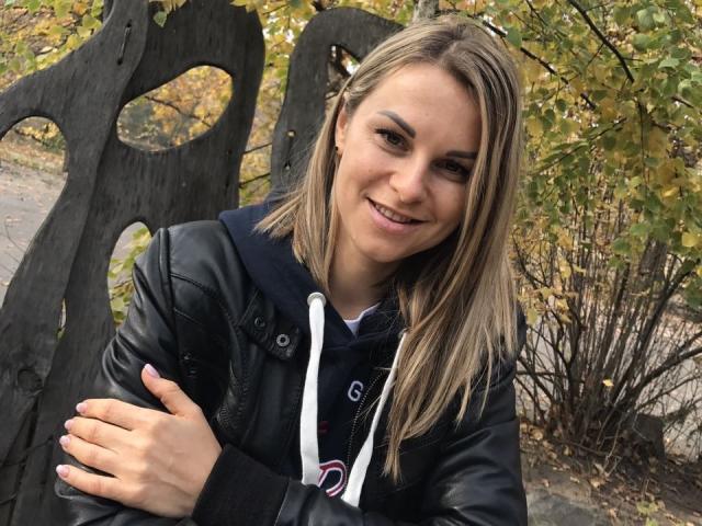 BelleLisaG avec des cheveux blonds, une veste en cuir noir et un sweat à capuche, souriante, les bras croisés, debout devant une sculpture en bois noir, avec des feuilles d'automne en arrière-plan..