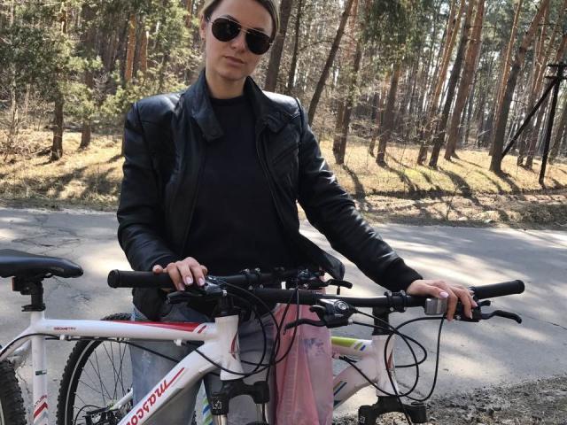 BelleLisaG, portant des lunettes de soleil, une veste en cuir noir et une jupe rose, se tient sur une route avec un vélo blanc, tenant le guidon. Elle se trouve dans une forêt avec de grands arbres..