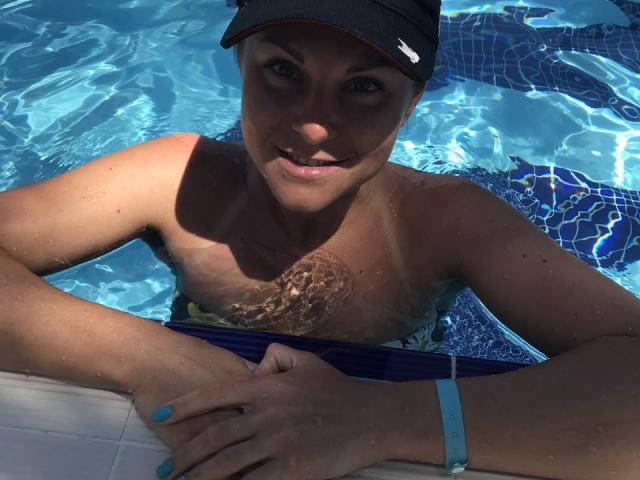 BelleLisaG dans une piscine, portant une casquette noire, un bracelet bleu et un maillot de bain à fleurs, avec de l'eau qui fronce sa poitrine et ses mains reposant sur le bord de la piscine..