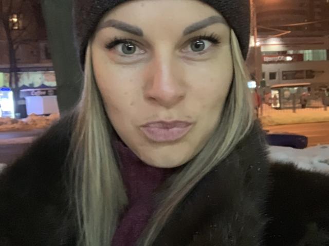 BelleLisaG a les lèvres pulpeuses, les yeux verts, les cheveux blonds, et porte un bonnet noir et un manteau en fourrure. De la neige est sur ses épaules. Elle a une expression mignonne et surprise..