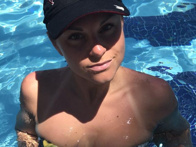 BelleLisaG, avec une peau bronzée et des yeux bleus, dans une piscine, portant une casquette noire, mouillée de gouttes d'eau, montrant ses seins nus, de petite à moyenne taille, regardant vers la caméra..