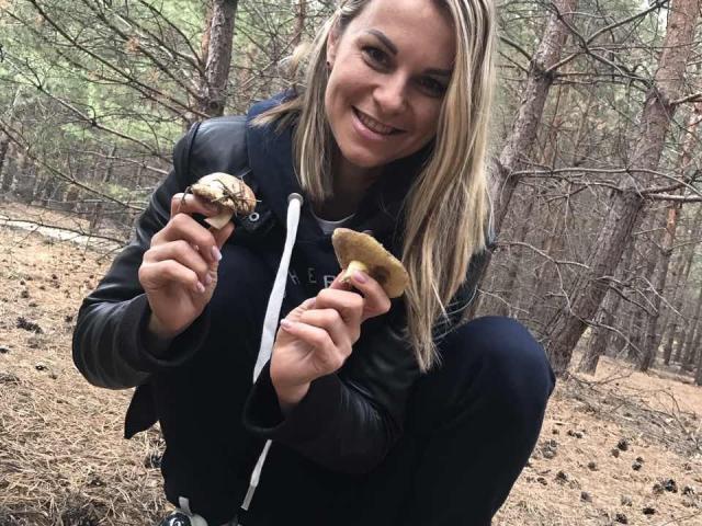 BelleLisaG, avec des cheveux blonds, souriante, tenant une noix de pin et un biscuit dans une forêt, portant une veste en cuir noir et un pantalon foncé, accroupie, montrant ses collations..