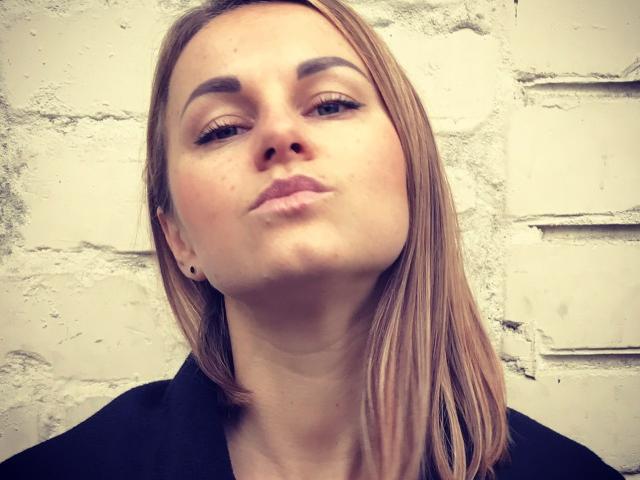 BelleLisaG avec des lèvres pulpeuses, des cheveux bruns clairs et lisses, une tenue noire, un maquillage discret et un sourcil légèrement levé, regardant légèrement vers le haut. Son nez est centré, et un petit boucle d'oreille est visible..