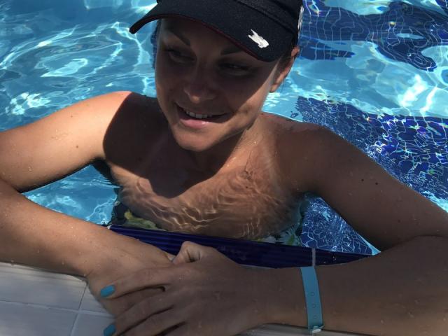 BelleLisaG dans une piscine, nue sur le buste, portant une casquette noire, un maillot de bain bleu et un bracelet bleu, souriante, les mains sur le bord de la piscine, des ondulations d'eau sur sa poitrine..