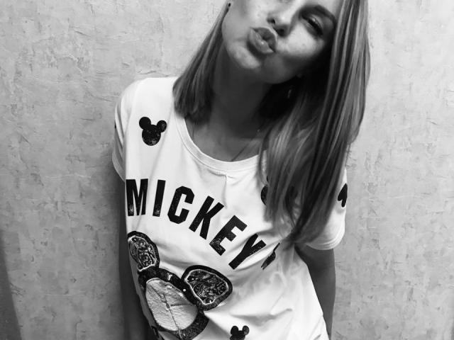 BelleLisaG, avec ses lèvres pulpeuses, appuyée contre un mur texturé, portant une chemise blanche "Mickey" avec un adorable motif de singe, des oreilles noires et des cheveux blonds lisses. Photo en noir et blanc..