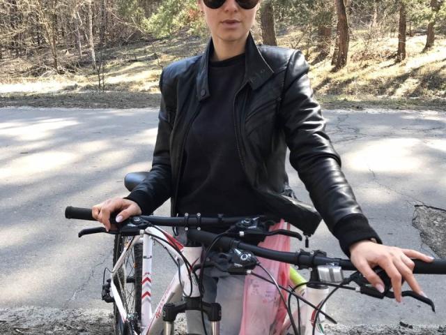 BelleLisaG en veste en cuir noir, haut noir et pantalon rose/gris, debout les mains sur le vélo, portant des lunettes de soleil, dans une forêt ensoleillée, avec un vélo aux accents rouge/blanc devant elle..