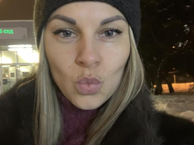 BelleLisaG, avec ses lèvres pulpeuses, fait une moue de baiser. Elle a les cheveux blonds, un bonnet gris, et porte un manteau en fourrure sombre avec une écharpe rouge..