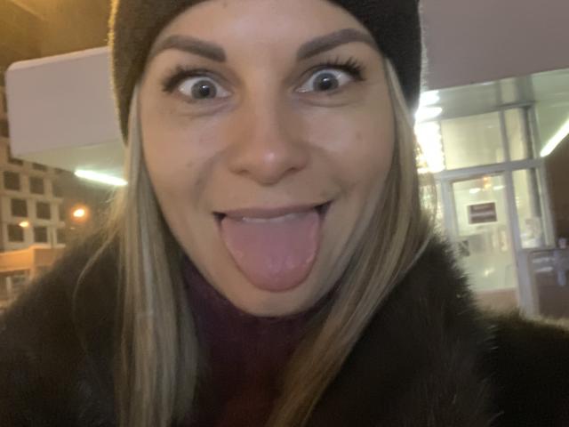 BelleLisaG, avec de grands yeux et la langue tirée, portant un bonnet noir et un manteau en fourrure, a les cheveux blonds et affiche une expression amusante et espiègle..