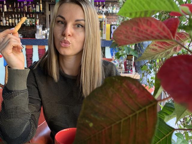 BelleLisaG avec des cheveux blonds, des lèvres pincées, un pull rayé, tenant un petit aliment, assise devant des feuilles rouges et vertes. Arrière-plan avec un bar flou..