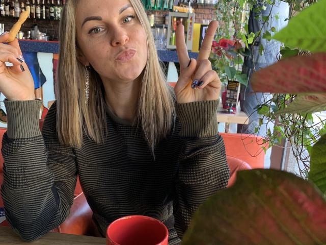 BelleLisaG avec des cheveux blonds, des lèvres gonflées et des doigts levés, portant un pull rayé, tenant un hot-dog, assise dans une cabine rouge, avec une tasse rouge devant elle..