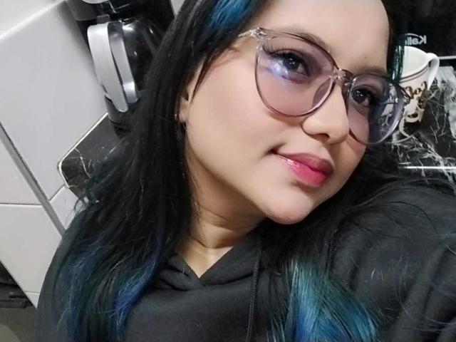 A AzulMoon tem cabelos pretos com pontas azuis, usa óculos e batom rosa, veste um moletom preto, tem um sorriso adorável e um rosto levemente corpulento, em uma selfie..