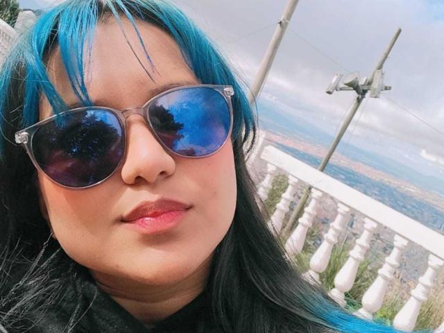 AzulMoon, com cabelo azul e preto, usando óculos de sol espelhados, tem lábios rosados e um leve sorriso. O fundo inclui um parapeito branco e um poste com luzes..