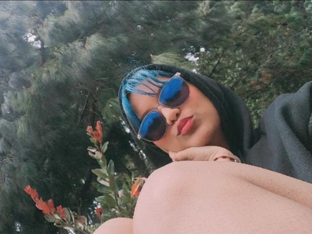 AzulMoon, con cabello azul, sudadera con capucha negra y gafas de sol reflectantes, labios fruncidos, rodilla levantada, un pino verde en el fondo. Luz suave, entorno natural, pose casual..