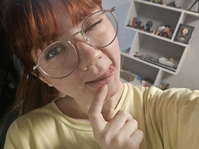 AlfrediaGhiloni with red hair, glasses, winking, and finger touching her lip, wearing a yellow shirt, showing a cute and playful expression.