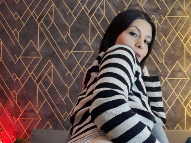 KarlaMazuera with black and white striped shirt, looking over shoulder, showing her ass, wearing red lipstick, dark hair, and gold eye makeup, in a modern, geometric room.