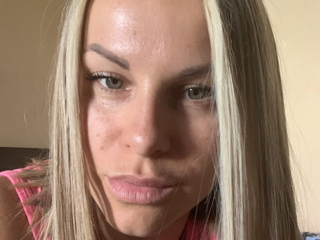 BelleLisaG a les cheveux blonds, les yeux verts, et un visage légèrement brillant, marqué par des cicatrices d'acné, avec des lèvres roses. Elle porte un haut rose et regarde droit devant la caméra avec une expression neutre..