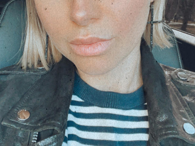 Le visage de BelleLisaG, avec sa peau pâle, ses lèvres roses et ses cheveux blonds, porte une veste en cuir vert par-dessus une chemise rayée bleu-vert, et elle a un anneau de nez du côté droit..